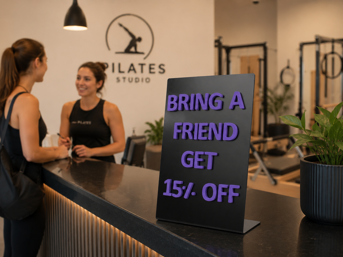Pilates stüdyosu resepsiyon bankosunda kullanılan mor yazılı metal kampanya panosu. Arka planda konuşan iki kişi ve pilates ekipmanları bulunan modern spor salonu ortamı.